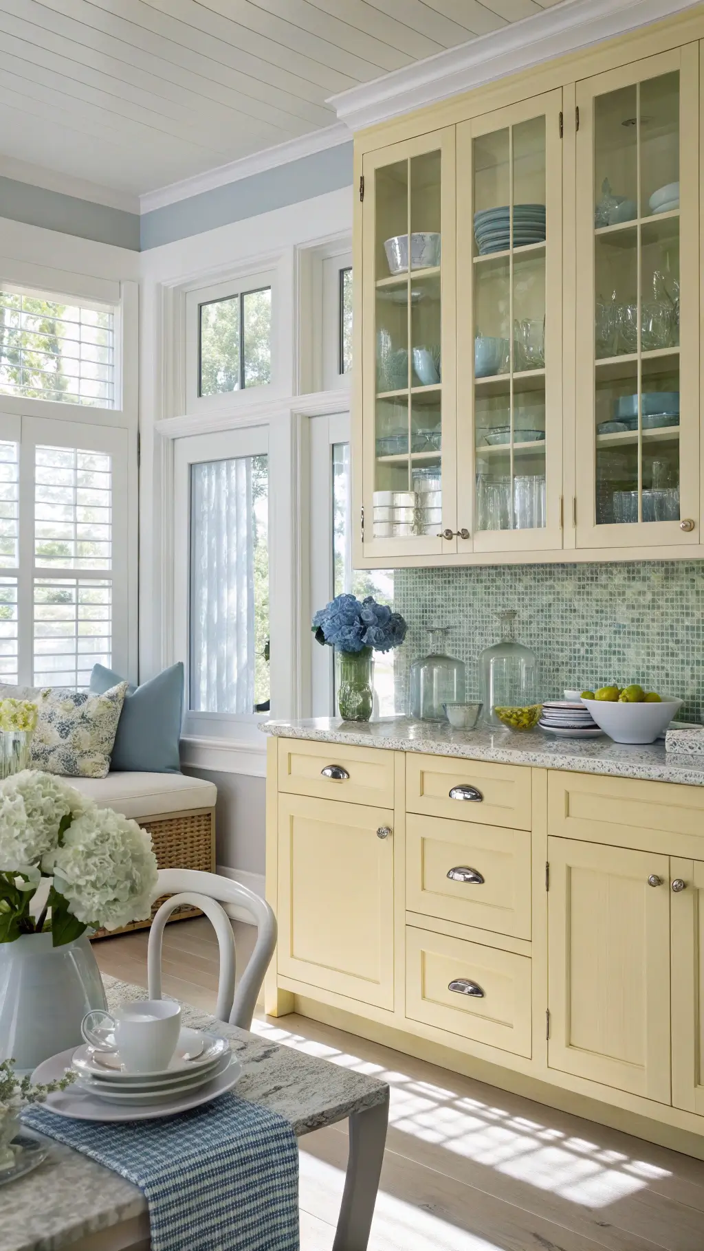 Sunny coastal-inspired kitchen with lemon beadboard cabinets, sea-glass mosaic backsplash, white shell handles, blue-grey accents, and fresh hydrangeas in a white pitcher