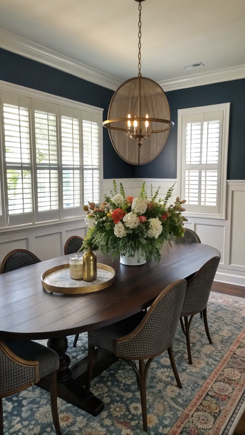 Transitional dining room with navy accent wall, wainscoting, dark oak oval table with a mixed media flower centerpiece in brass vessels, bathed in golden hour light filtering through plantation shutters.