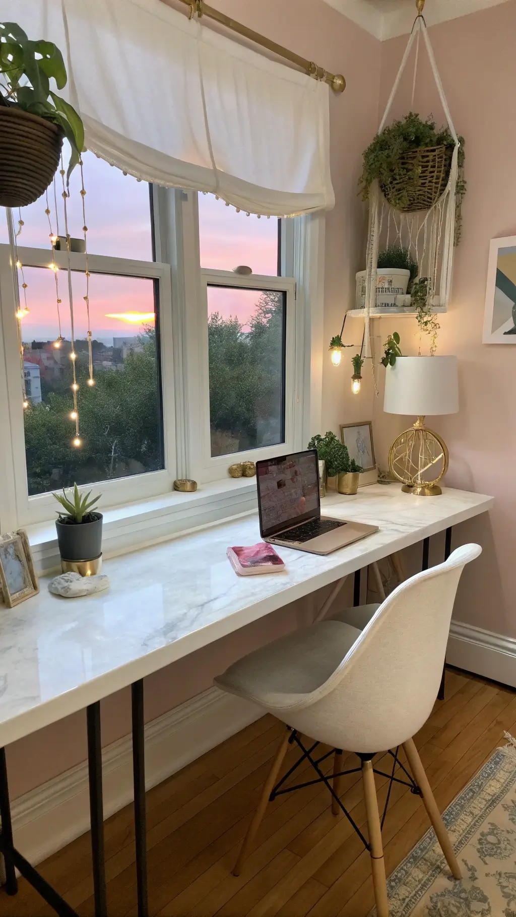 Feminine dusk-lit workspace with white marble desk on a windowsill, ghost chair, linen roman shade, string of pearls plants, blush pink accent wall and brass accessories.