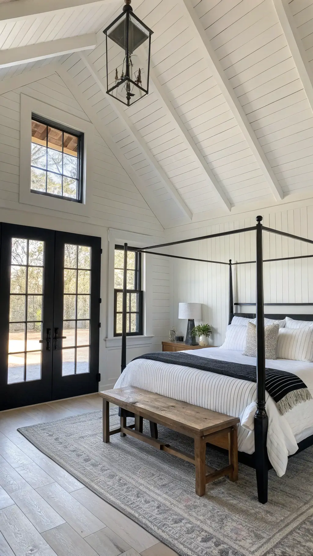 Modern farmhouse bedroom featuring white shiplap walls, black French doors, an iron canopy bed with white linens, black wooden bench, ceramic table lamps on raw wood nightstands, and cathedral ceiling lit by morning light