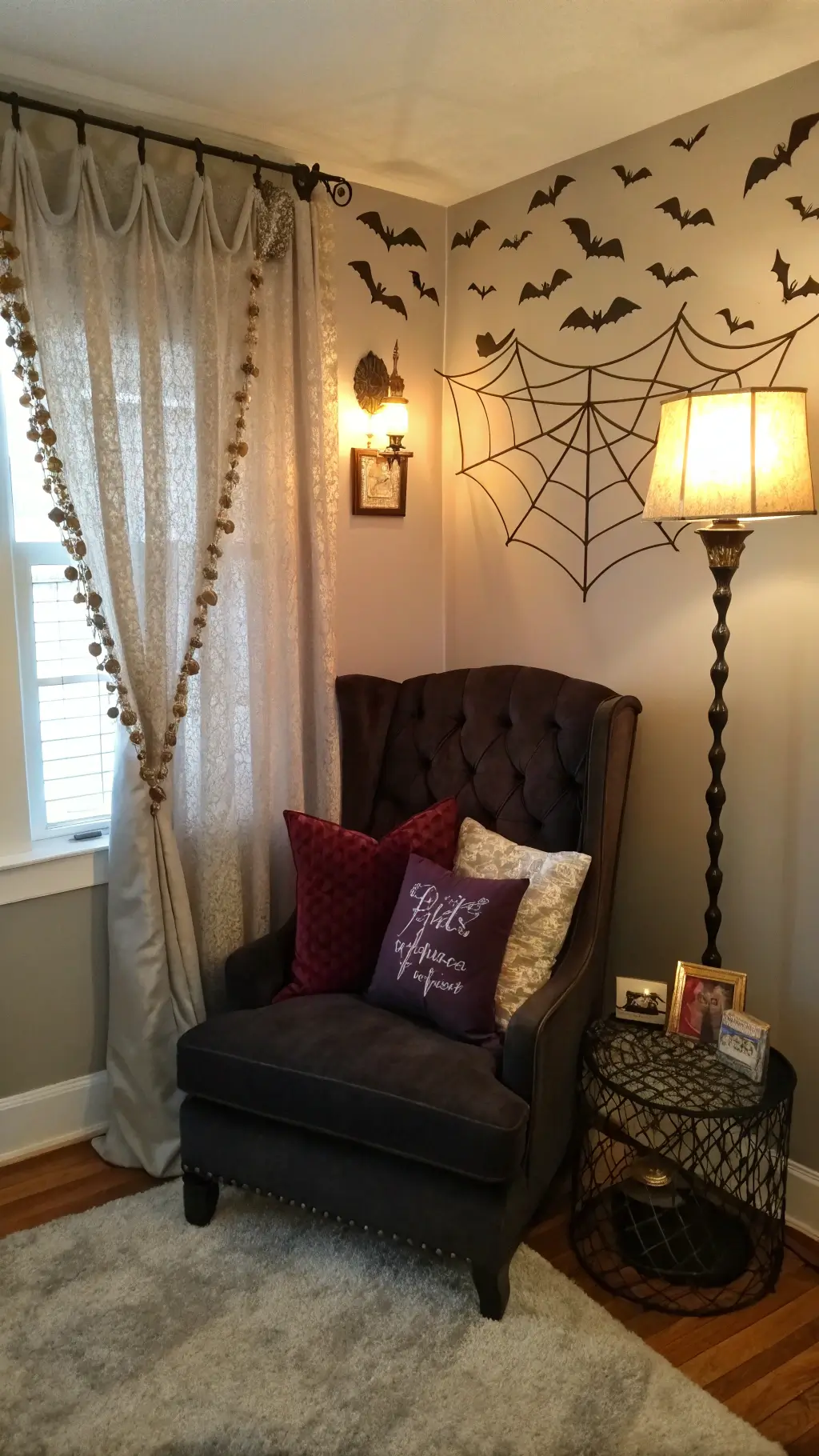 Gothic-style reading nook in master bedroom with oversized black velvet wingback chair, cascading paper bat installation, vintage brass lamp casting spiderweb shadows, and burgundy and silver throw pillows, lit by golden hour light through sheer curtains.